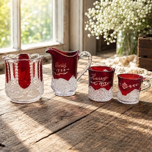 Peut inclure: Un ensemble de quatre pièces en verre rouge rubis et transparent avec des motifs taillés complexes. La collection comprend un bol large, un pichet gravé "Doris 1929", une petite tasse gravée "Harry 1917" et une petite tasse à anse avec gravure florale.