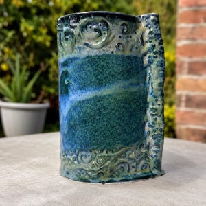 Peut inclure: Un vase cylindrique en céramique avec un émail tacheté bleu profond et vert. La surface est ornée de motifs en spirale en relief en haut et en bas, et une colonne verticale texturée de bosses en relief longe le côté droit.