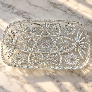 May include: A rectangular clear glass serving tray featuring an intricate pressed pattern of multiple starburst motifs and geometric shapes. The edges of the tray are scalloped and faceted, catching the light to create a brilliant reflective effect.