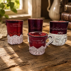 Puede incluir: Cuatro objetos de recuerdo de vidrio rojo rubí con bases de vidrio prensado transparente que presentan intrincados patrones geométricos. Una pequeña taza con asa tiene la inscripción Souvenir of Uxbridge, Ontario. Otras piezas incluyen textos como Port Rowan, Ontario, Hamilton y Mother-in-law 1893.