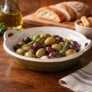 May include: A ceramic bowl filled with green and dark red olives, garnished with rosemary sprigs. The bowl sits on a wooden table, with a loaf of bread and olive oil in the background. A rustic food presentation.