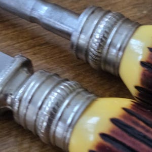 May include: Close-up of two knife handles. The handles have a yellow and brown pattern with a silver metal bolster. The blades are silver and the background is a wooden surface.