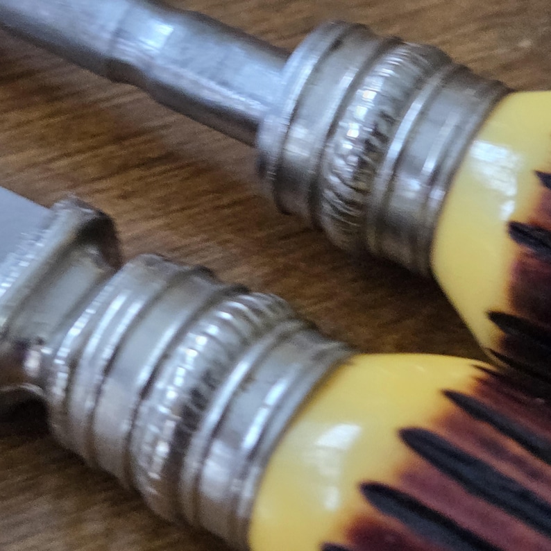 May include: Close-up of two knife handles. The handles have a yellow and brown pattern with a silver metal bolster. The blades are silver and the background is a wooden surface.
