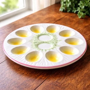 Puede incluir: Plato blanco de cerámica para servir huevos rellenos con borde rosa y diseño floral verde en el centro. El plato tiene 10 hendiduras en forma de huevo, cada una llena de una sustancia amarilla. El plato está sobre una superficie de madera.