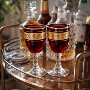 May include: Two red wine glasses with gold trim and a decorative gold band featuring a floral design. The glasses are on a gold bar cart with liquor bottles in the background. The glasses are filled with a red liquid.