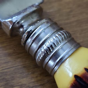 May include: Close-up of a butter knife with a silver metal handle and a yellow and brown handle. The handle has a decorative silver band with a rope-like design. The blade is silver and slightly curved.
