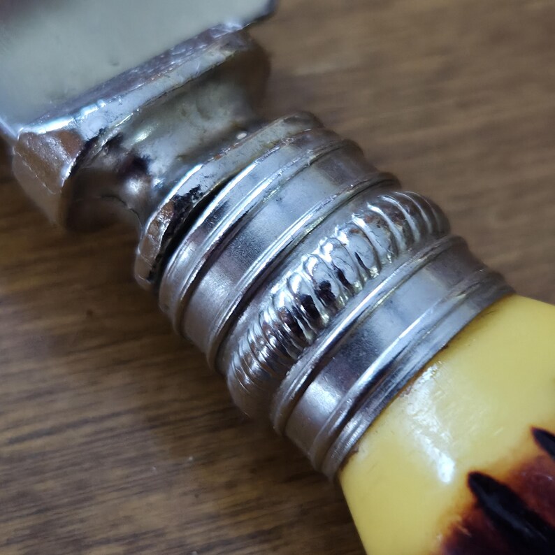 May include: Close-up of a butter knife with a silver metal handle and a yellow and brown handle. The handle has a decorative silver band with a rope-like design. The blade is silver and slightly curved.