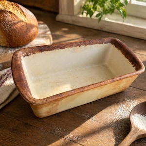 May include: A rectangular ceramic loaf tin with a cream interior and a distressed brown rim. The tin is on a wooden surface, suggesting a rustic aesthetic. The tin is empty, ready for baking. A loaf of bread is visible in the background.
