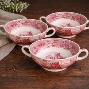 May include: Three ceramic soup bowls with handles, featuring a red and white pastoral scene design. The bowls are arranged on a dark wooden surface, showcasing their classic, vintage aesthetic. The bowls are ideal for serving soup or other dishes.
