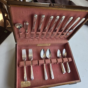 May include: A wooden cutlery case with a pink velvet lining, open to reveal an assortment of silver utensils. The set includes knives, forks, and spoons with ornate handles. The case is labelled "Community" and "Conti-Tarnish".