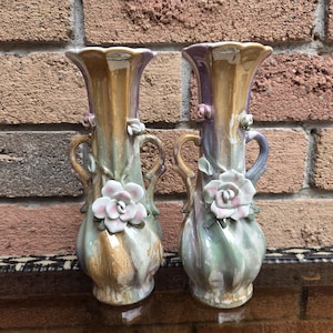 May include: A pair of iridescent ceramic vases featuring a swirling pastel glaze of gold, lavender, and sage green. Each vase has two side handles and is decorated with three-dimensional sculpted ceramic flowers in white and soft pink.