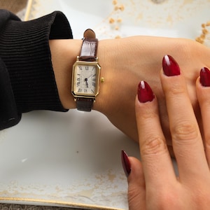 May include: A gold rectangular wristwatch with a brown leather band is displayed. The watch face is white with black Roman numerals. The watch is on a wrist.