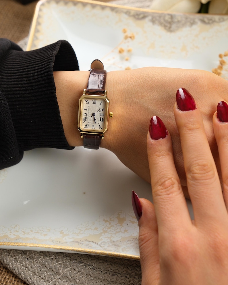May include: A gold rectangular wristwatch with a brown leather band is displayed. The watch face is white with black Roman numerals. The watch is on a wrist.