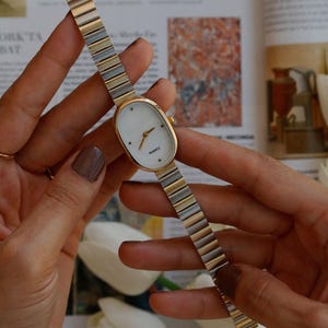 May include: A gold-toned quartz watch with a white oval face and a gold and silver band. The watch is held by a person. The watch face has gold hands and the word "QUARTZ" printed on it. The band is made of small, rectangular links.