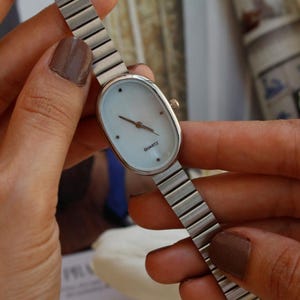 May include: A silver-toned quartz watch with an oval face and a light-colored dial. The watch has a silver band and is held by a person. The word "QUARTZ" is printed on the watch face.