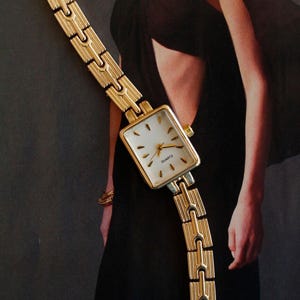 Op de afbeelding: Goudkleurig quartz horloge met een rechthoekige witte wijzerplaat en gouden wijzers. De horlogeband is een bijpassende gouden schakelstijl. Het horloge wordt gepresenteerd tegen een donkere achtergrond, wat wijst op een mode- of accessoirethema.
