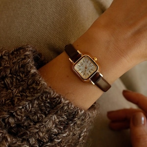 Puede incluir: Un reloj cuadrado dorado con esfera blanca y números negros. El reloj tiene una correa de cuero marrón. El reloj se lleva en la muñeca, con una manga de suéter marrón y beige en primer plano.