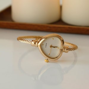 May include: Gold-toned heart-shaped watch with a white face and gold hands. The watch has a twisted metal band. The word "Quartz" is printed on the watch face. The watch is displayed on a white surface.