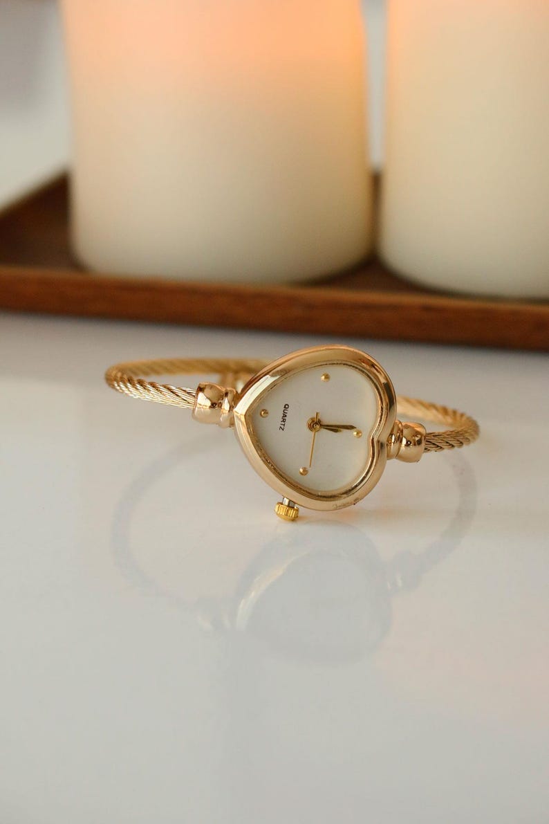 May include: Gold-toned heart-shaped watch with a white face and gold hands. The watch has a twisted metal band. The word "Quartz" is printed on the watch face. The watch is displayed on a white surface.