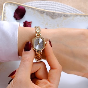 May include: A gold-toned wristwatch featuring a hexagonal face with a silver dial and simple hour markers. The watch band is constructed from interconnected gold-toned metal links, creating a chain-like appearance that secures around the wrist.