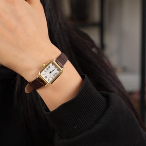 May include: A gold rectangular wristwatch with a white face and Roman numeral hour markers. The watch has a brown leather band. The watch is on a wrist, and the person is wearing a black long-sleeve shirt.