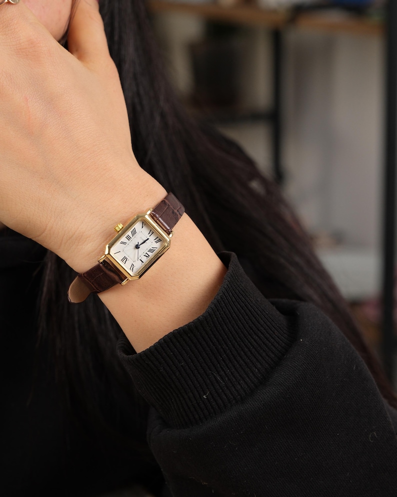 May include: A gold rectangular wristwatch with a white face and Roman numeral hour markers. The watch has a brown leather band. The watch is on a wrist, and the person is wearing a black long-sleeve shirt.