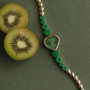 May include: A gold-tone wristwatch featuring a heart-shaped case with a green dial and a bracelet composed of alternating metallic gold-tone and solid green heart-shaped links. The watch is positioned next to two sliced kiwi fruit halves.