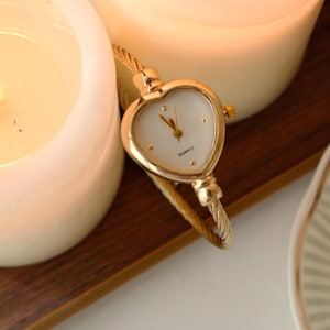 Peut inclure: Une montre à quartz dorée avec un cadran en forme de cœur et un bracelet tressé. La montre est posée près de deux bougies blanches allumées sur un plateau en bois. Le cadran de la montre est blanc avec des aiguilles et des index dorés.