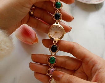 May include: A gold-toned wristwatch with a series of colorful, oval-shaped gemstones. The watch face is round with a light-colored dial. The gemstones include green, black, and pink stones. The watch is held by a person.
