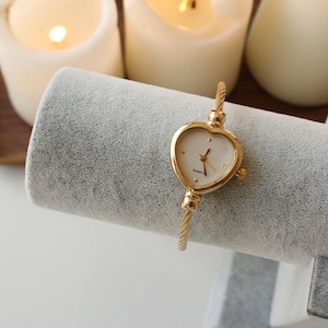 May include: A gold-toned heart-shaped watch with a white face and delicate gold band. The watch is displayed on a gray velvet jewelry stand, with lit candles in the background. The watch is a stylish accessory.