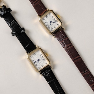 May include: Two rectangular gold-toned watches with white faces and Roman numeral hour markers. One watch has a black leather band, the other a brown leather band. The watch faces have blue hands and the word "QUARTZ" is printed on the face.