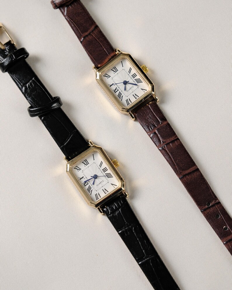 May include: Two rectangular gold-toned watches with white faces and Roman numeral hour markers. One watch has a black leather band, the other a brown leather band. The watch faces have blue hands and the word "QUARTZ" is printed on the face.