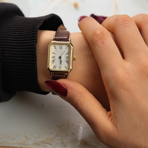 May include: A gold rectangular wristwatch with a white face and Roman numeral hour markers. The watch has a brown leather band. The watch is on a wrist.