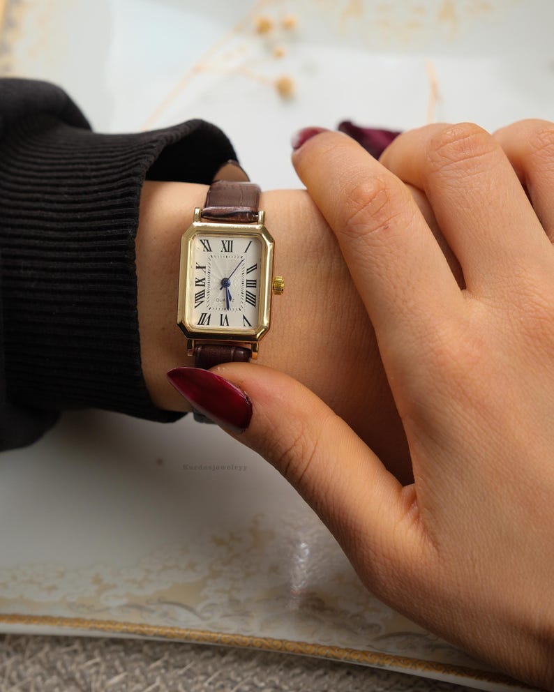 May include: A gold rectangular wristwatch with a white face and Roman numeral hour markers. The watch has a brown leather band. The watch is on a wrist.
