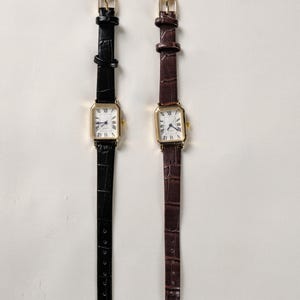 May include: Two rectangular gold-toned watches with white faces and Roman numeral hour markers. One watch has a black leather band, the other a brown leather band. Both have gold buckles.