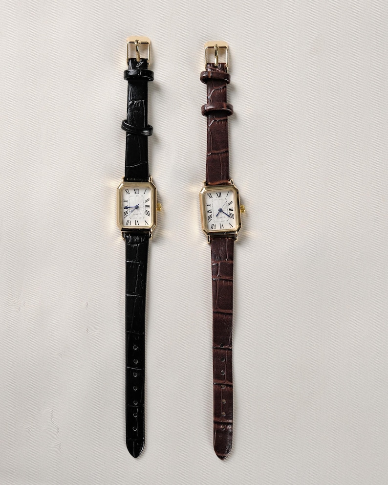 May include: Two rectangular gold-toned watches with white faces and Roman numeral hour markers. One watch has a black leather band, the other a brown leather band. Both have gold buckles.