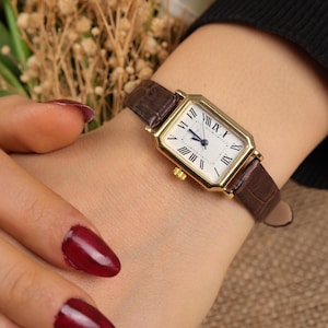 May include: A gold rectangular wristwatch with a white face and black Roman numerals. The watch has a brown leather band. The watch is a classic design.