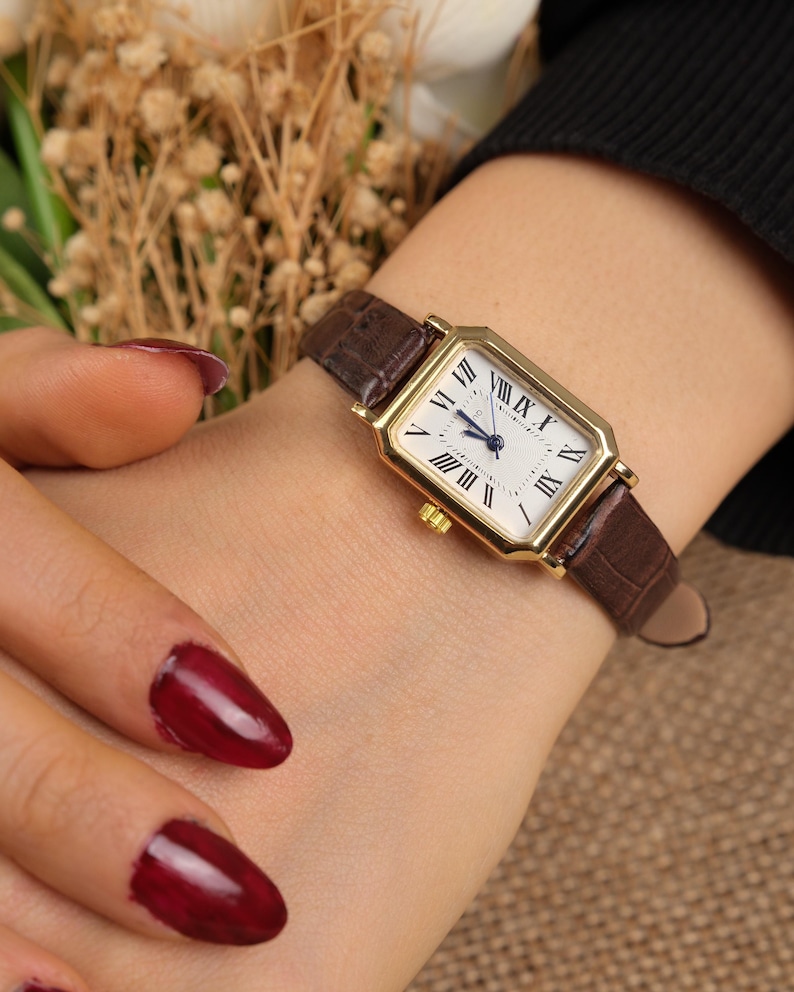 May include: A gold rectangular wristwatch with a white face and black Roman numerals. The watch has a brown leather band. The watch is a classic design.