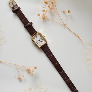 May include: A rectangular gold-toned wristwatch with a white face and black Roman numerals. The watch has a brown leather band with a crocodile pattern and a gold buckle. The watch is displayed on a white surface with dried flowers.