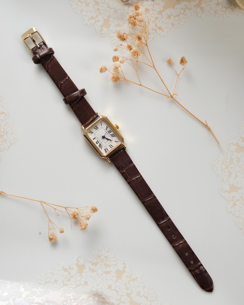 May include: A rectangular gold-toned wristwatch with a white face and black Roman numerals. The watch has a brown leather band with a crocodile pattern and a gold buckle. The watch is displayed on a white surface with dried flowers.
