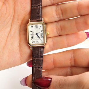 May include: A gold-toned rectangular watch with a white face and Roman numeral hour markers. The watch has a brown leather band with a crocodile pattern. The watch is a classic style.