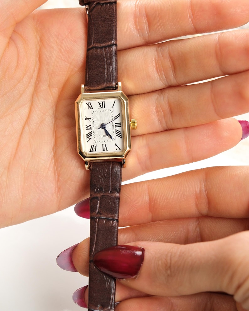 May include: A gold-toned rectangular watch with a white face and Roman numeral hour markers. The watch has a brown leather band with a crocodile pattern. The watch is a classic style.