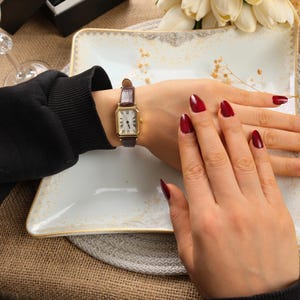 May include: A gold rectangular wristwatch with a brown leather band. The watch face has black Roman numerals and hands. The person's hand is resting on a white plate with a gold trim. The nails are painted a dark red.