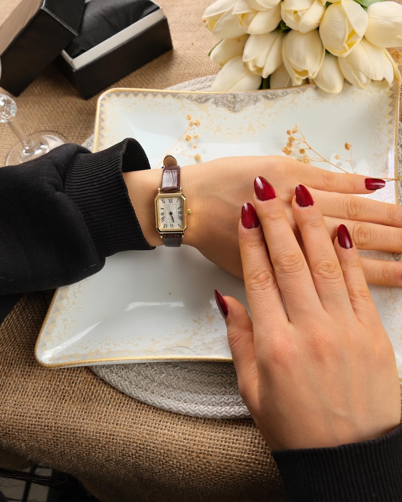 May include: A gold rectangular wristwatch with a brown leather band. The watch face has black Roman numerals and hands. The person's hand is resting on a white plate with a gold trim. The nails are painted a dark red.