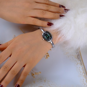 May include: A silver-toned bangle watch with an oval, dark green face and Roman numeral hour markers. The watch is on a wrist, with a white fluffy background. The watch band is a thin, curved design.