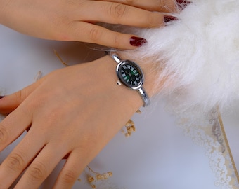 Könnte beinhalten: Eine silberfarbene Armbanduhr mit einem ovalen, dunkelgrünen Zifferblatt und römischen Ziffern. Die Uhr befindet sich an einem Handgelenk, vor einem weißen, flauschigen Hintergrund. Das Armband ist ein dünnes, gebogenes Design.