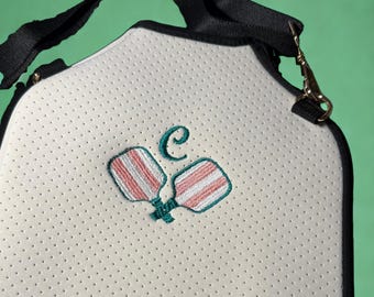 May include: White pickleball paddle bag with a black strap and trim. The bag features a perforated texture and an embroidered design of two pickleball paddles with red and white stripes and a teal letter "C".