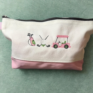 May include: White and pink golf-themed cosmetic bag with a black zipper. The bag features embroidered images of golf clubs, a golf ball, a golf cart, and a golf bag. The bag is made of canvas material.