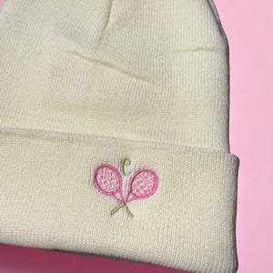 May include: Cream-colored beanie hat with a folded brim. The hat features an embroidered design of two pink tennis rackets with green stems and a small letter "C". The hat is made of a soft, knitted material.