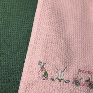 May include: A green golf towel with a black trim and a pink golf towel with embroidered golf-themed images. The pink towel features a golf bag, golf ball, clubs, and a golf cart. Both towels have a textured waffle weave.
